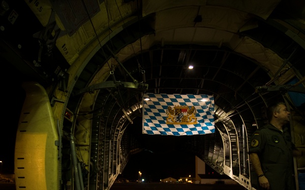 The image shows the interior of a large military transport aircraft, likely a C-130 Hercules, photographed at night with its rear cargo ramp open. A blue and white diamond-patterned flag with a coat of arms — identifiable as the flag of Bavaria (Free State of Bavaria, Germany) — is hanging from the aircraft's ceiling. A uniformed crew member stands on the right side near the open ramp, looking outward onto what appears to be a nighttime airfield.