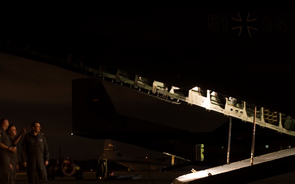 A nighttime scene on an airport tarmac showing a large military transport aircraft with its rear cargo ramp lowered and illuminated. Two or three personnel in flight suits are standing near the aircraft, apparently in conversation. A German military insignia (Iron Cross) is partially visible on the fuselage, suggesting this is a German Air Force (Luftwaffe) aircraft.