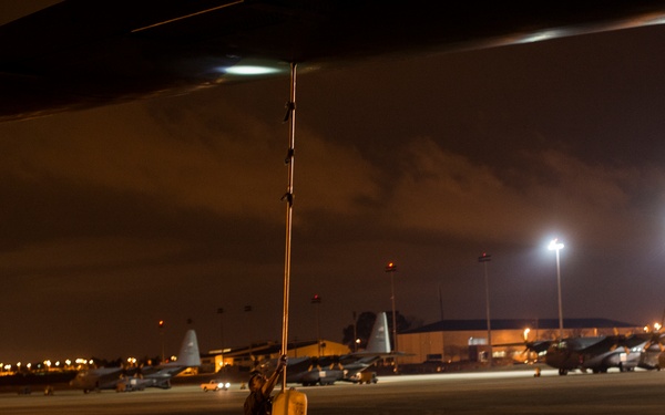 A nighttime scene on a military flight line showing a ground crew member standing beneath a large aircraft wing, holding an extended pole with a container attached. Multiple military transport aircraft, likely C-130 Hercules, are visible parked on the tarmac in the background. The scene is illuminated by airport floodlights and ambient ramp lighting.