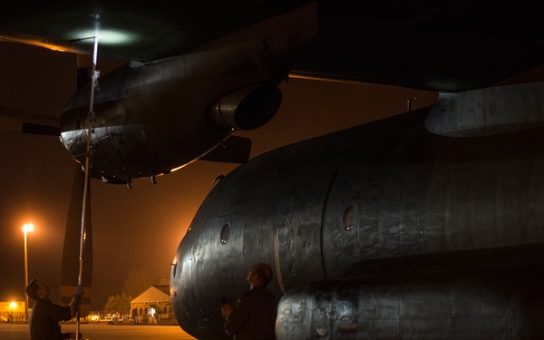 A nighttime maintenance or pre-flight inspection scene on a military airfield tarmac. Two personnel in flight suits are conducting work on a large military aircraft, likely a C-130 Hercules or similar turboprop transport, illuminated by ambient ramp lighting. The low-light conditions suggest either late-night or early-morning operations.