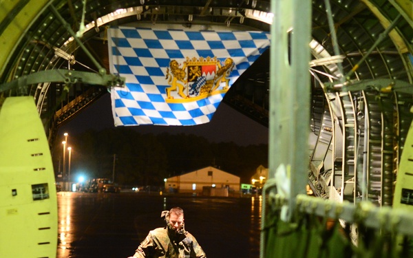 The image is taken from inside a large military cargo aircraft, looking out through the open rear cargo ramp toward a wet tarmac at night. A person in military/flight gear is walking up the ramp carrying a red object, while another crew member is visible on the right side wearing a headset. A blue and white Bavarian state flag (Freistaat Bayern) is prominently displayed above the aircraft opening.