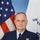 An official portrait photograph of a senior U.S. Coast Guard officer in dress uniform, seated at a table with hands clasped. The nameplate on his uniform reads 'COLLINS' and 'U.S. COAST GUARD'. He is flanked by the American flag and what appears to be a U.S. Coast Guard or departmental flag.
