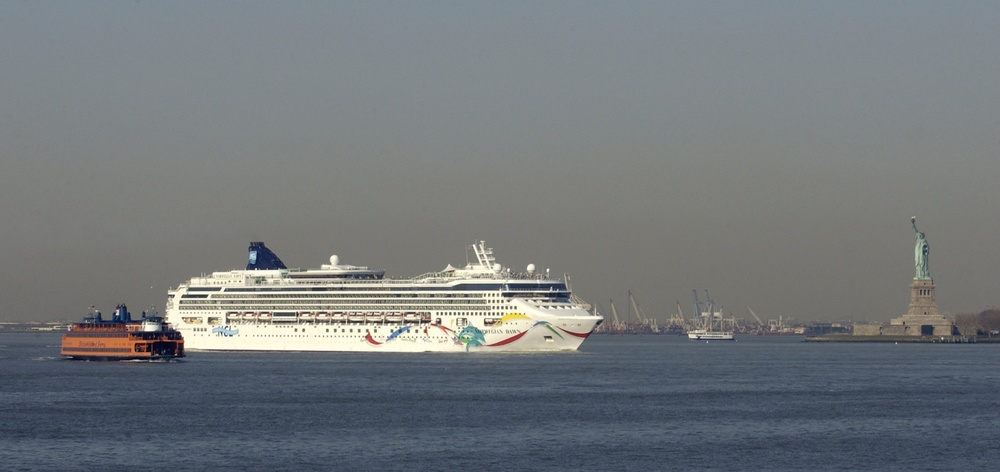 A large Norwegian Cruise Line ship, identifiable as the Norwegian Dawn, navigates New York Harbor with the Statue of Liberty visible in the background on the right. A Staten Island Ferry is visible to the left of the cruise ship. The scene captures a classic New York Harbor view with industrial port infrastructure visible in the middle distance.