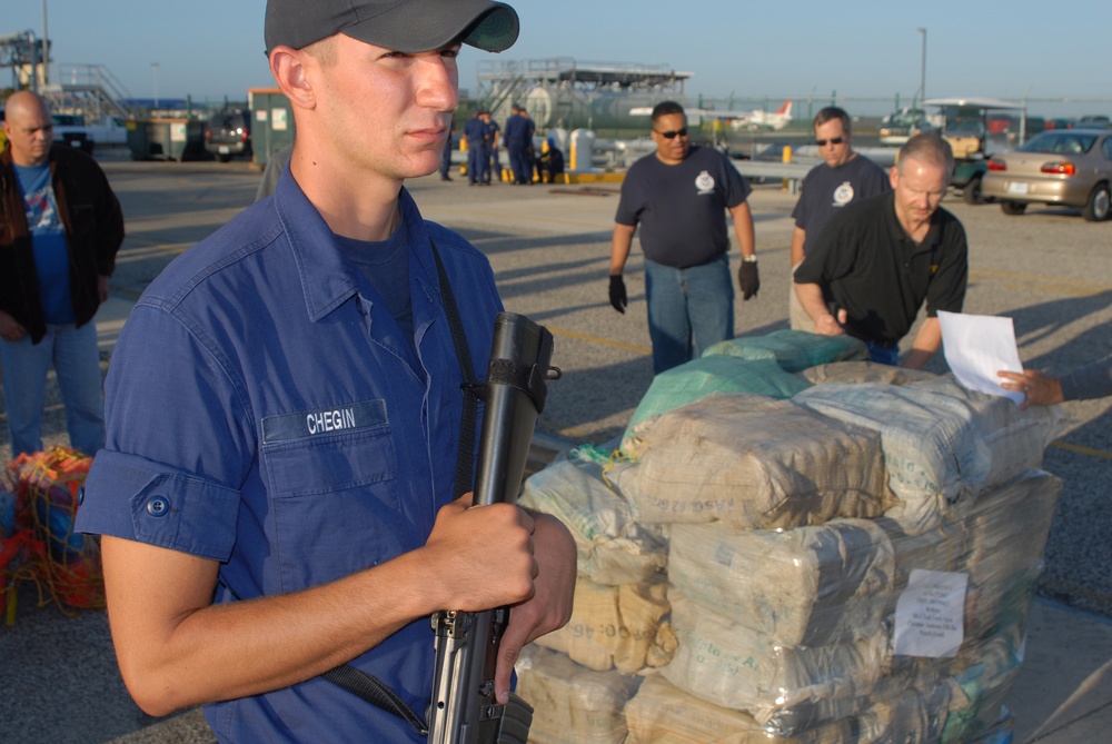 A U.S. Coast Guard member named Chegin stands guard with a rifle next to a large pallet of seized bundled packages, likely narcotics, at what appears to be a port or airfield facility. Other personnel, including individuals in law enforcement attire, are examining documents near the seized contraband. This appears to be an official drug interdiction offload or evidence processing event.