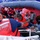 A U.S. Coast Guard rescue operation is underway on open water, with personnel assisting civilians aboard an inflatable rigid-hull inflatable boat (RHIB). Coast Guard members in red life vests and blue uniforms are helping at least two civilians, including a woman wearing an orange life jacket. The scene depicts an active maritime rescue or interdiction operation.