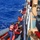 U.S. Coast Guard personnel are conducting a maritime rescue operation, transferring individuals from an orange rigid inflatable boat (RIB) onto a larger Coast Guard vessel. Multiple Coast Guard crew members in red life vests and helmets are assisting civilians, who appear to be migrants or distressed mariners, up a ladder or gangway onto the ship. The operation is taking place in open blue ocean waters.
