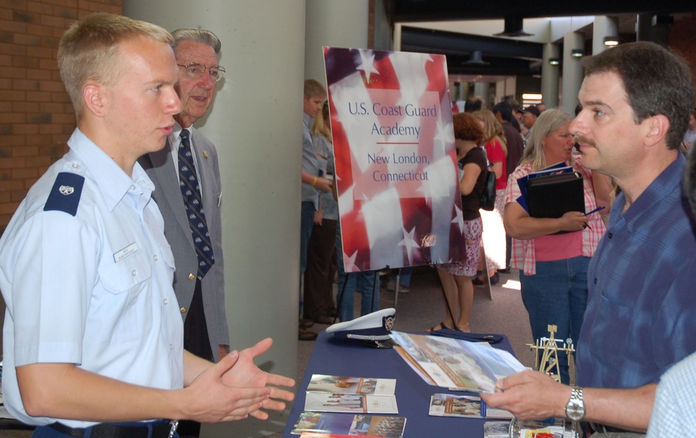 A U.S. Coast Guard Academy recruitment or information fair booth is shown, located in New London, Connecticut. A young cadet in uniform is speaking with a civilian man across a table covered with brochures and informational materials. The event appears to be a college or military academy fair held in a large indoor venue.