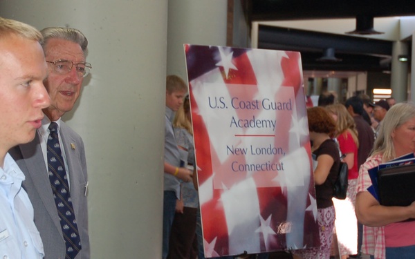 A U.S. Coast Guard Academy recruitment or information fair booth is shown, located in New London, Connecticut. A young cadet in uniform is speaking with a civilian man across a table covered with brochures and informational materials. The event appears to be a college or military academy fair held in a large indoor venue.