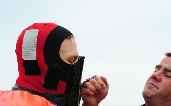 Two individuals are shown in what appears to be a maritime safety training or preparation scenario. One person is wearing an orange immersion/survival suit with a red and black hood, while the other, wearing a navy blue uniform and red life vest, is assisting by zipping up the hood of the survival suit. The setting appears to be outdoors on or near a watercraft under overcast skies.