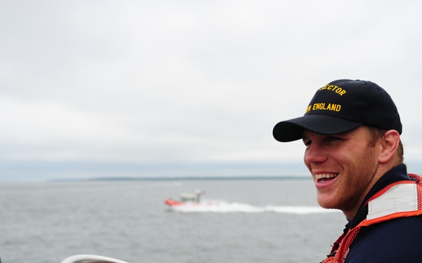 A smiling young man is piloting a boat on open water while wearing a red life vest and a dark navy cap with yellow text reading 'SECTOR NEW ENGLAND.' He is seated at the helm with a stainless steel steering wheel and marine navigation equipment visible to his left. In the background, another vessel can be seen moving at speed across the water.