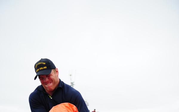 A U.S. Coast Guard member wearing a 'USCG Sector New England' cap is assisting someone in removing or donning an orange immersion/survival suit on the deck of a vessel. In the background, a U.S. Coast Guard patrol boat is visible on the water with personnel on deck. This appears to be a training exercise or post-rescue operation.