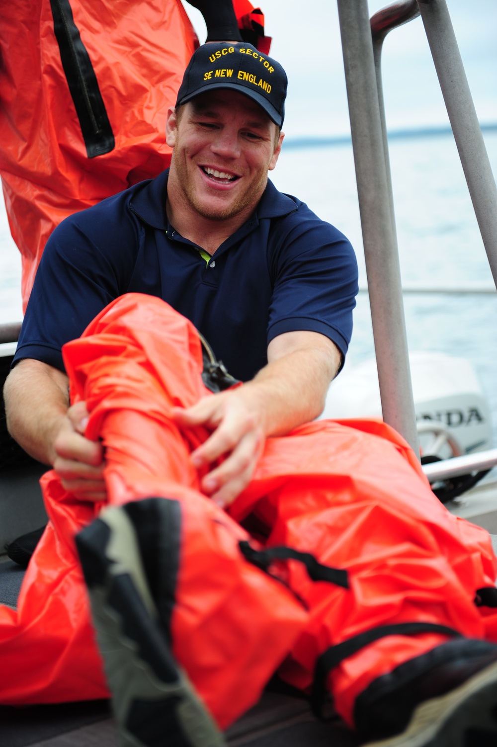 A smiling young man wearing a navy blue polo shirt and a cap reading 'USCG SECTOR SE NEW ENGLAND' is seated on what appears to be a boat. He is handling bright orange immersion/survival suits, likely preparing or stowing safety equipment. The setting is an open watercraft with metal railings and a Honda outboard motor visible in the background.