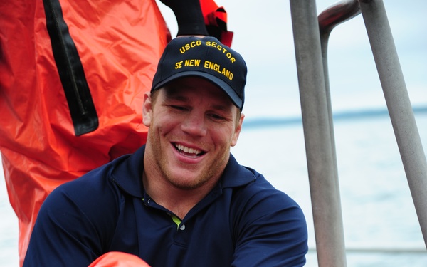 A smiling young man wearing a navy blue polo shirt and a cap reading 'USCG SECTOR SE NEW ENGLAND' is seated on what appears to be a boat. He is handling bright orange immersion/survival suits, likely preparing or stowing safety equipment. The setting is an open watercraft with metal railings and a Honda outboard motor visible in the background.