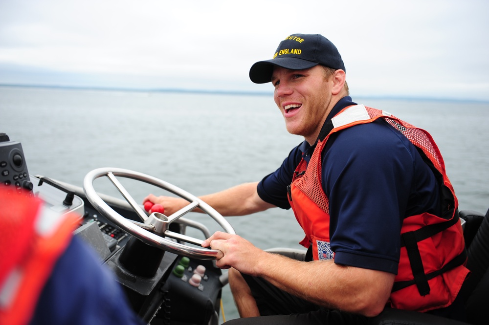 A young man is piloting a boat on open water while smiling broadly at the camera. He is wearing a navy blue uniform shirt, an orange life vest with a Coast Guard insignia, and a dark cap with yellow text reading 'SECTOR NEW ENGLAND.' The setting appears to be an overcast day on a calm body of water.