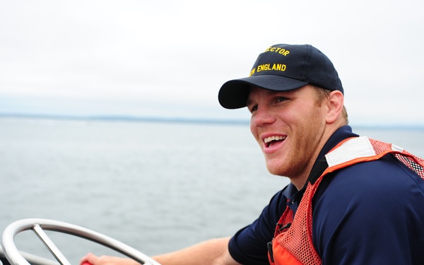 A young man is piloting a boat on open water while smiling broadly at the camera. He is wearing a navy blue uniform shirt, an orange life vest with a Coast Guard insignia, and a dark cap with yellow text reading 'SECTOR NEW ENGLAND.' The setting appears to be an overcast day on a calm body of water.
