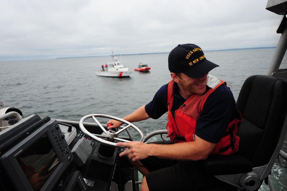 A U.S. Coast Guard member is piloting a patrol boat on open water, wearing an orange life vest and a cap reading 'USCG SECTOR SE NEW ENGLAND'. He is smiling while operating the helm with navigation equipment visible in the foreground. Two additional Coast Guard vessels are visible in the background on calm, overcast waters.
