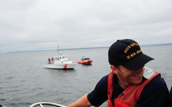 A U.S. Coast Guard member is piloting a patrol boat on open water, wearing an orange life vest and a cap reading 'USCG SECTOR SE NEW ENGLAND'. He is smiling while operating the helm with navigation equipment visible in the foreground. Two additional Coast Guard vessels are visible in the background on calm, overcast waters.