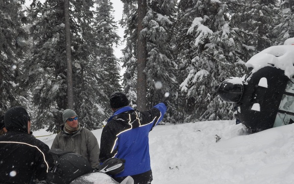 A group of people are gathered in a snowy, forested area around snowmobiles. A vehicle — appearing to be a truck or SUV — is visible overturned or heavily buried in a large snow bank on the right side of the image. Yellow caution/barrier tape is visible at the bottom right, suggesting this may be an accident or recovery scene.