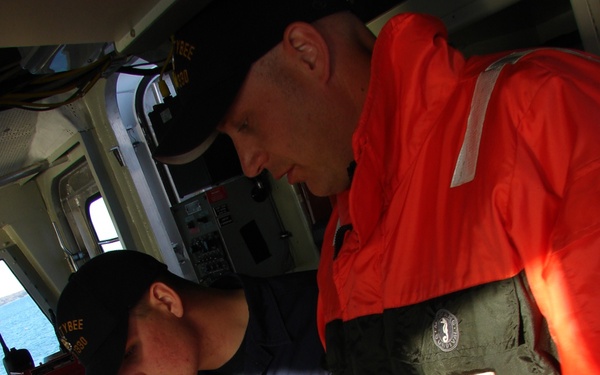 Two U.S. Coast Guard personnel are aboard a vessel's bridge or pilothouse, reviewing a nautical chart together. One crew member wears a blue U.S. Coast Guard uniform with a name tag reading 'WARDSON' and a cap marked 'TYBEE,' while the other wears an orange and dark green immersion/survival suit with a Coast Guard insignia patch. A third person is partially visible in the background near navigation equipment.