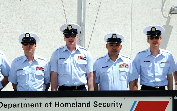 Six U.S. Coast Guard personnel in dress uniform are posing in front of an official sign for LORAN-C Station Fallon, NV, Master 9940, under the U.S. Department of Homeland Security. The group includes enlisted members and at least one officer (rightmost individual with gold insignia on cap and commander-level rank insignia). The photo appears to be an official unit or command photograph taken outdoors against a white building.