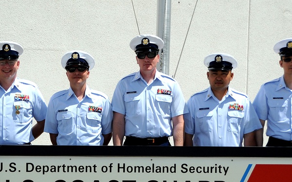 Five U.S. Coast Guard personnel in uniform are posed behind an official station sign at LORAN-C Station Fallon, NV, Master 9940. The sign identifies the facility as part of the U.S. Department of Homeland Security, U.S. Coast Guard. The personnel are wearing light blue short-sleeve uniforms with white covers (caps) and various ribbons/medals.