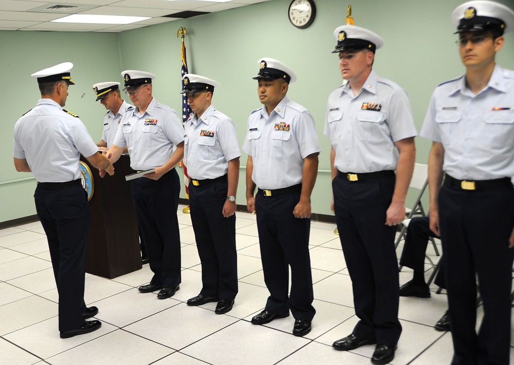 A formal military ceremony is taking place indoors, likely an awards or recognition ceremony. A senior officer on the left is presenting a document or certificate to personnel standing in a line. The uniformed individuals appear to be members of the United States Coast Guard based on their distinctive uniforms and insignia.