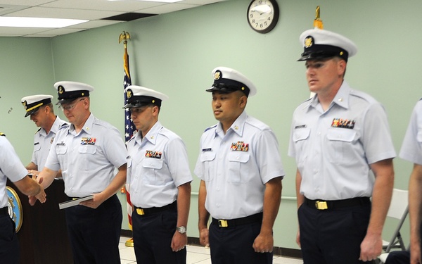 A formal military ceremony is taking place indoors, likely an awards or recognition ceremony. A senior officer on the left is presenting a document or certificate to personnel standing in a line. The uniformed individuals appear to be members of the United States Coast Guard based on their distinctive uniforms and insignia.