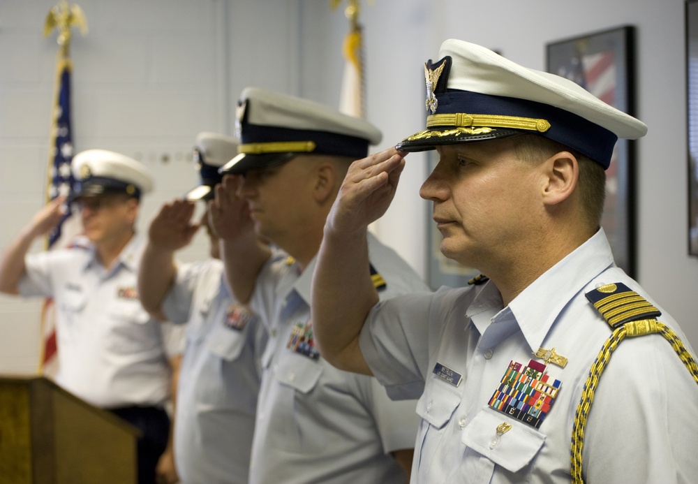 Three U.S. Coast Guard officers are standing in a line and rendering a salute during what appears to be a formal military ceremony. The officers are dressed in white service uniforms with rank insignia, ribbons, and cover caps. An American flag and what appears to be a Coast Guard or unit flag are visible in the background.