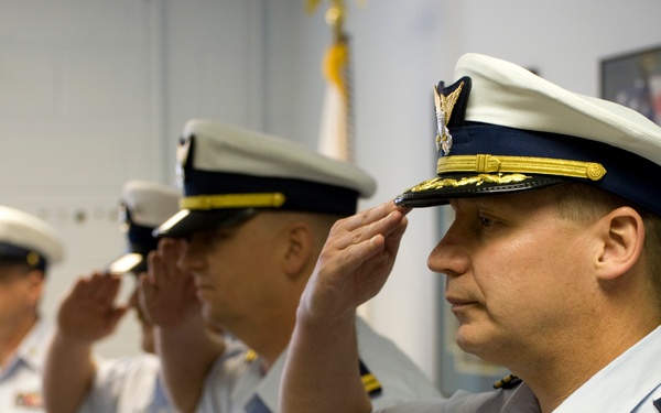 Three U.S. Coast Guard officers are standing in a line and rendering a salute during what appears to be a formal military ceremony. The officers are dressed in white service uniforms with rank insignia, ribbons, and cover caps. An American flag and what appears to be a Coast Guard or unit flag are visible in the background.