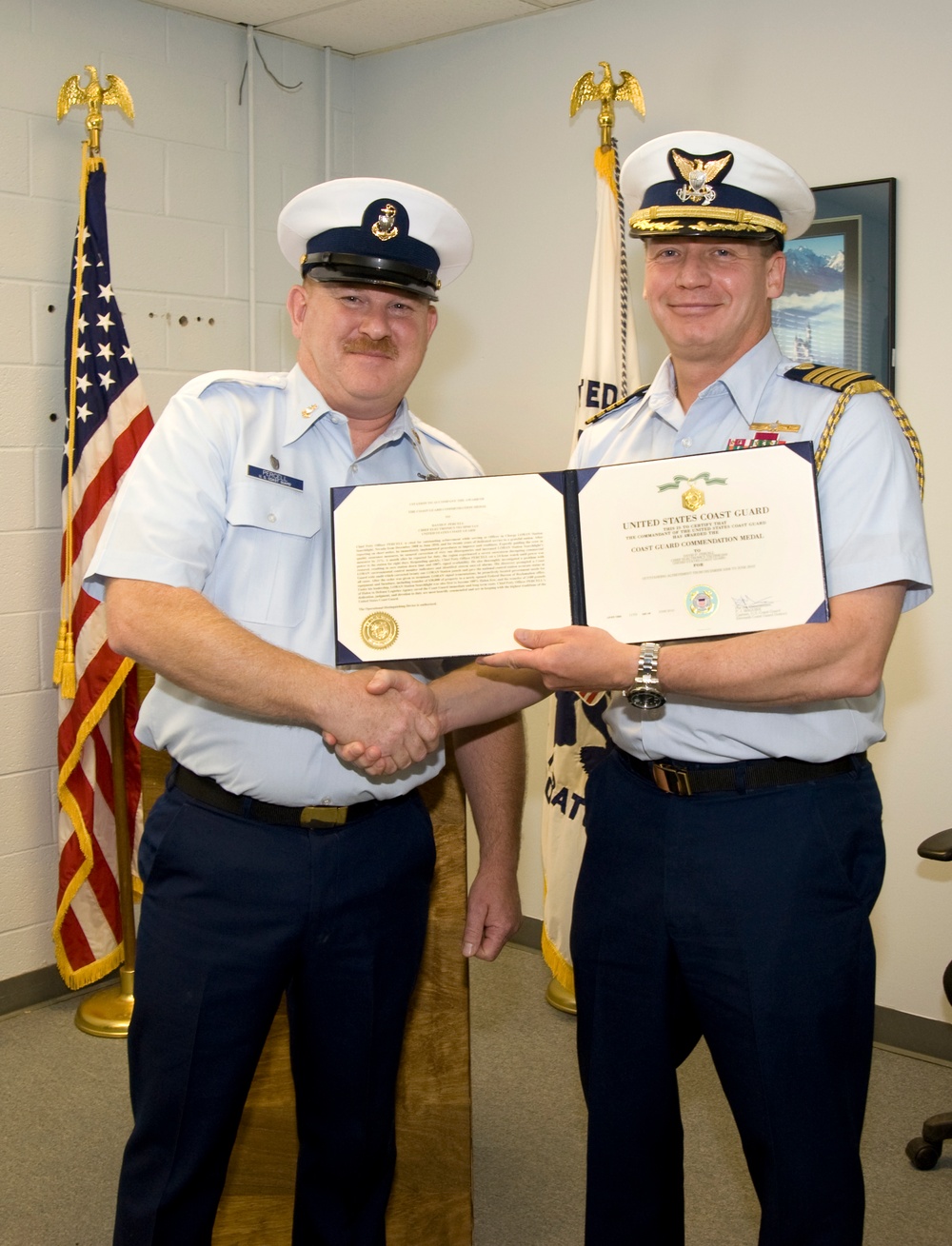 Two United States Coast Guard personnel are posing together while holding an open certificate folder displaying a 'Coast Guard Commendation Medal' award. The officer on the right, wearing a higher-ranking uniform with gold braid and ribbons, is shaking hands with the enlisted member on the left in a formal award presentation ceremony. An American flag and a Coast Guard flag are visible in the background.