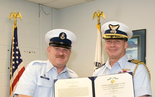 Two United States Coast Guard personnel are posing together while holding an open certificate folder displaying a 'Coast Guard Commendation Medal' award. The officer on the right, wearing a higher-ranking uniform with gold braid and ribbons, is shaking hands with the enlisted member on the left in a formal award presentation ceremony. An American flag and a Coast Guard flag are visible in the background.