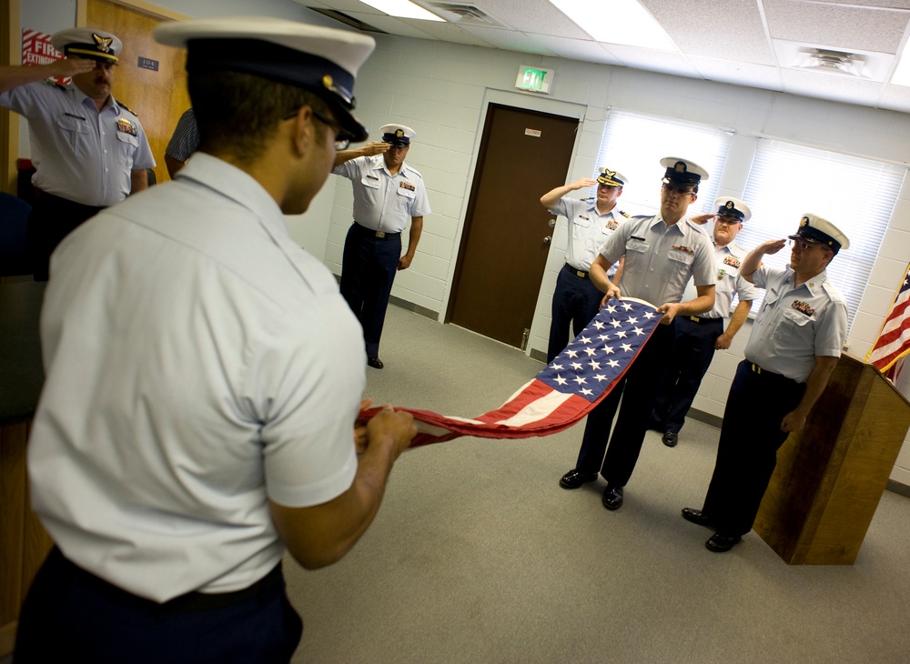 A group of uniformed military or Coast Guard personnel are conducting a formal flag folding ceremony inside an indoor facility. Several members are saluting while two individuals hold and fold an American flag. The setting appears to be a meeting room or administrative building.