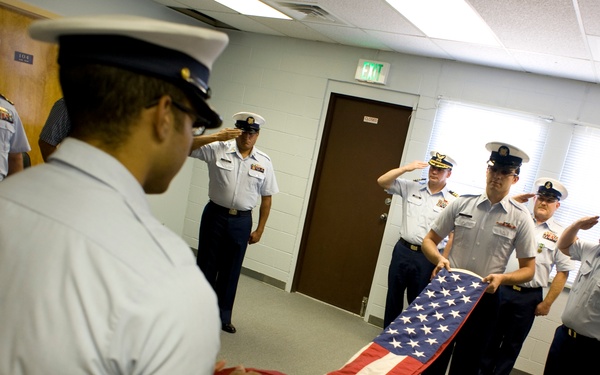 A group of uniformed military or Coast Guard personnel are conducting a formal flag folding ceremony inside an indoor facility. Several members are saluting while two individuals hold and fold an American flag. The setting appears to be a meeting room or administrative building.