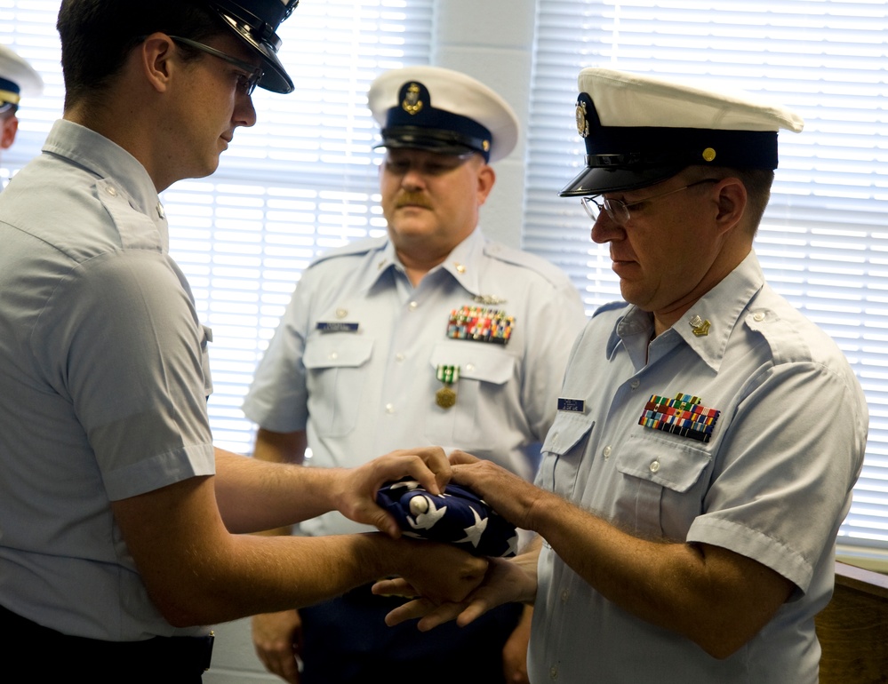 A formal military ceremony is taking place in which a folded American flag is being transferred between two uniformed personnel, likely a Coast Guard retirement or memorial ceremony. Three individuals in U.S. Coast Guard uniforms are visible, with the central action being the presentation of a tri-folded flag. The setting appears to be an indoor official space with window blinds visible in the background.