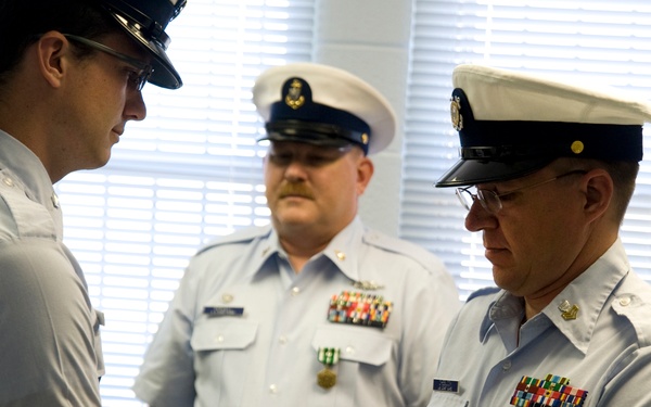 A formal military ceremony is taking place in which a folded American flag is being transferred between two uniformed personnel, likely a Coast Guard retirement or memorial ceremony. Three individuals in U.S. Coast Guard uniforms are visible, with the central action being the presentation of a tri-folded flag. The setting appears to be an indoor official space with window blinds visible in the background.
