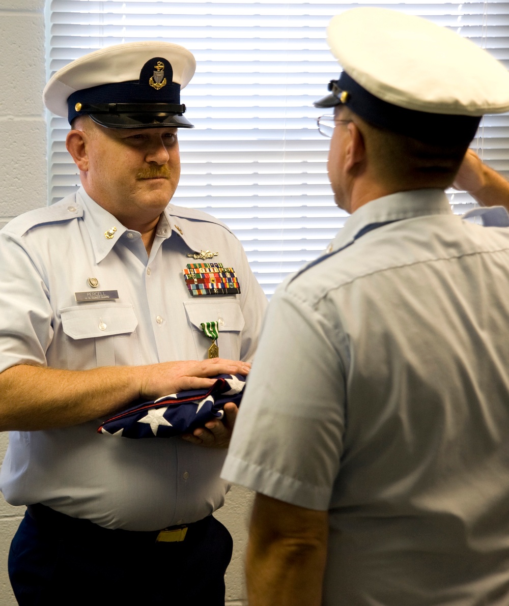 Two U.S. Coast Guard personnel are engaged in a formal military ceremony, likely a retirement or award presentation. The service member facing the camera, identified by nameplate as 'PERCELL,' is holding a folded American flag and wearing dress uniform with multiple ribbons and medals. The second individual, shown from behind, appears to be rendering a salute.