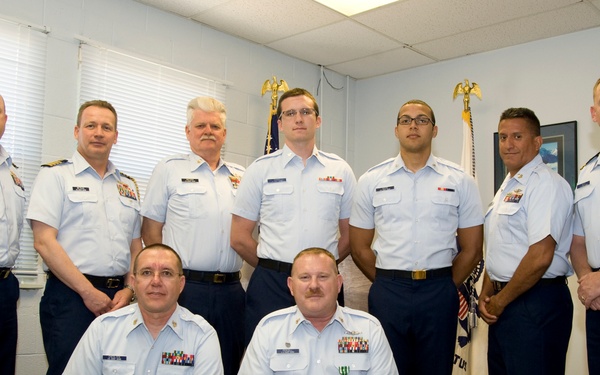 A formal group photograph of nine United States Coast Guard personnel in a military office or administrative setting. Two individuals are kneeling in the front row while seven stand in the back row. One person in the front row is wearing a medal, suggesting this may be an award or recognition ceremony.