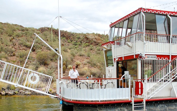 A two-deck riverboat or lake cruise vessel, registered in Arizona (AZ 1432AE), is docked near a rocky desert shoreline with saguaro cacti visible on the hillside. The white and red boat appears to be a tourist or excursion vessel, with a gangway/boarding ramp extended toward the shore. At least two crew members are visible on the vessel, and a mooring rope extends from the bow into the water.