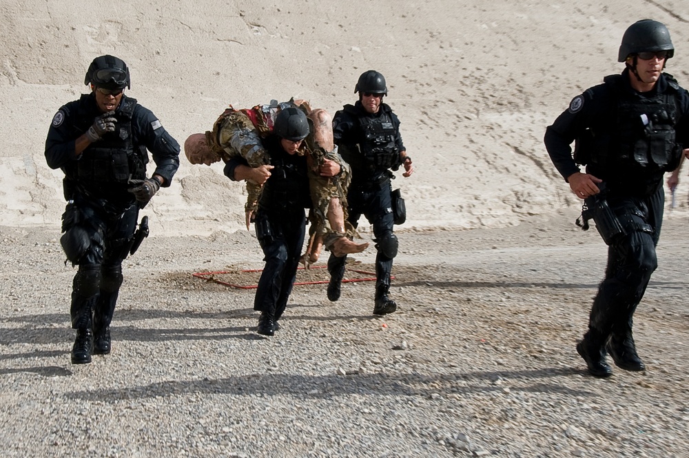 The image shows four individuals dressed in black tactical gear and helmets running across a gravel/dirt surface near a light-colored concrete or stone wall. One officer is carrying an incapacitated person wearing camouflage clothing over their shoulder in a fireman's carry. This appears to be a tactical training exercise or emergency response drill.