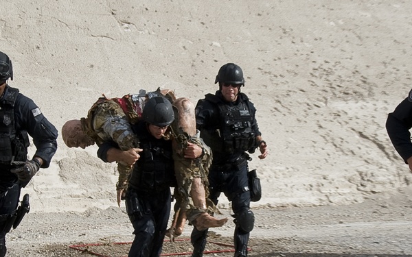 The image shows four individuals dressed in black tactical gear and helmets running across a gravel/dirt surface near a light-colored concrete or stone wall. One officer is carrying an incapacitated person wearing camouflage clothing over their shoulder in a fireman's carry. This appears to be a tactical training exercise or emergency response drill.