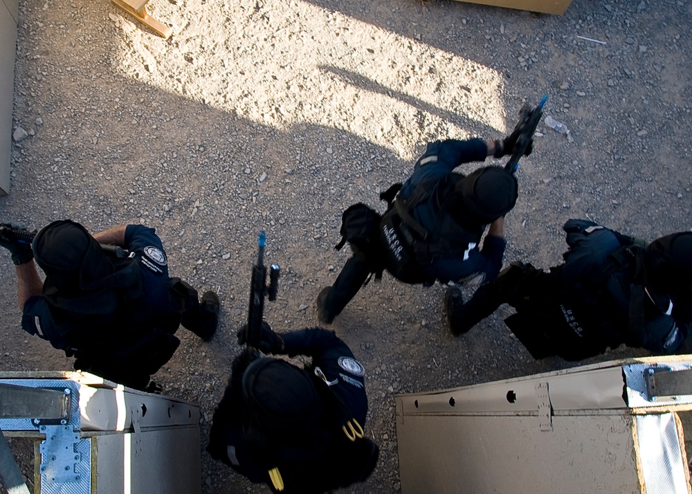 An aerial/overhead photograph showing four tactical personnel dressed in black uniforms and gear moving in formation across a gravel/dirt surface. The image appears to depict a law enforcement or military training exercise, with personnel carrying weapons and wearing body armor. The photo is taken from directly above, likely from an elevated position or rooftop.