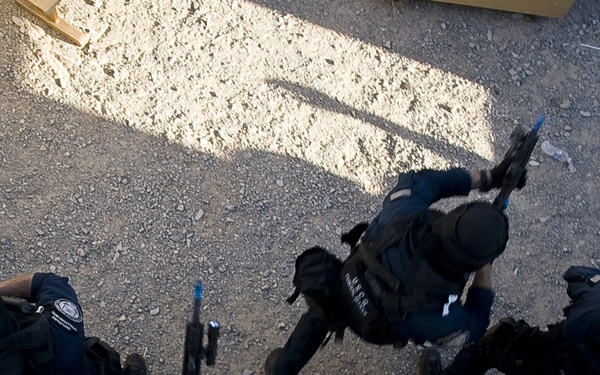 An aerial/overhead photograph showing four tactical personnel dressed in black uniforms and gear moving in formation across a gravel/dirt surface. The image appears to depict a law enforcement or military training exercise, with personnel carrying weapons and wearing body armor. The photo is taken from directly above, likely from an elevated position or rooftop.