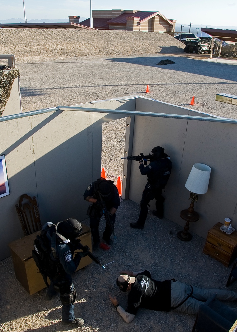 The image shows a tactical training exercise taking place in an outdoor mock room or building structure, viewed from an elevated angle. Three personnel in tactical gear and helmets are conducting a room-clearing drill, with one person on the ground appearing to play the role of a subject or detainee. The setting includes training props such as furniture, orange traffic cones, and a gravel surface consistent with a military or law enforcement training facility.