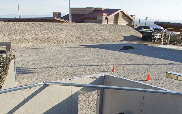 The image shows a tactical training exercise taking place in an outdoor mock room or building structure, viewed from an elevated angle. Three personnel in tactical gear and helmets are conducting a room-clearing drill, with one person on the ground appearing to play the role of a subject or detainee. The setting includes training props such as furniture, orange traffic cones, and a gravel surface consistent with a military or law enforcement training facility.