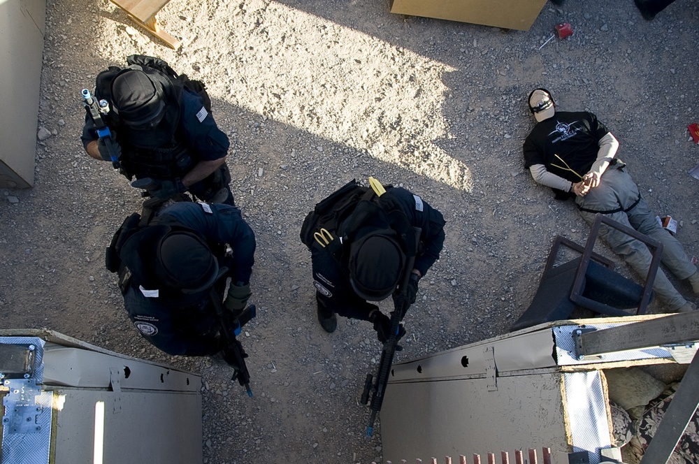 An aerial/overhead photograph showing three tactical personnel in black uniforms and body armor surrounding a restrained individual on the ground. The scene appears to be a law enforcement or military training exercise, with one person lying on gravel with hands bound behind their back. The image is taken from an elevated vantage point, possibly from a vehicle or structure.