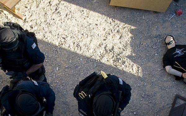 An aerial/overhead photograph showing three tactical personnel in black uniforms and body armor surrounding a restrained individual on the ground. The scene appears to be a law enforcement or military training exercise, with one person lying on gravel with hands bound behind their back. The image is taken from an elevated vantage point, possibly from a vehicle or structure.