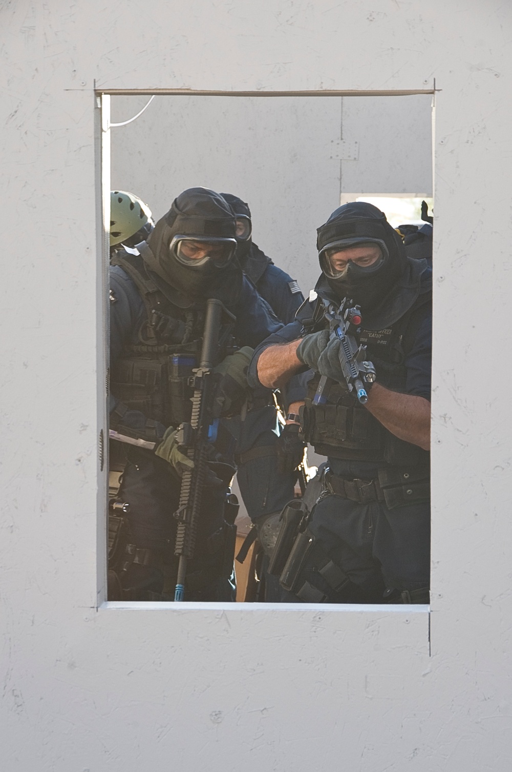 The image shows tactical law enforcement or military personnel viewed through a rectangular window opening in a white wall. Multiple operators are visible wearing full tactical gear including balaclavas, protective goggles, body armor, and tactical vests while holding rifles. The scene appears to be a training exercise or operational scenario involving building entry or room clearing.