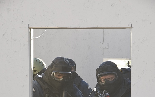 The image shows tactical law enforcement or military personnel viewed through a rectangular window opening in a white wall. Multiple operators are visible wearing full tactical gear including balaclavas, protective goggles, body armor, and tactical vests while holding rifles. The scene appears to be a training exercise or operational scenario involving building entry or room clearing.
