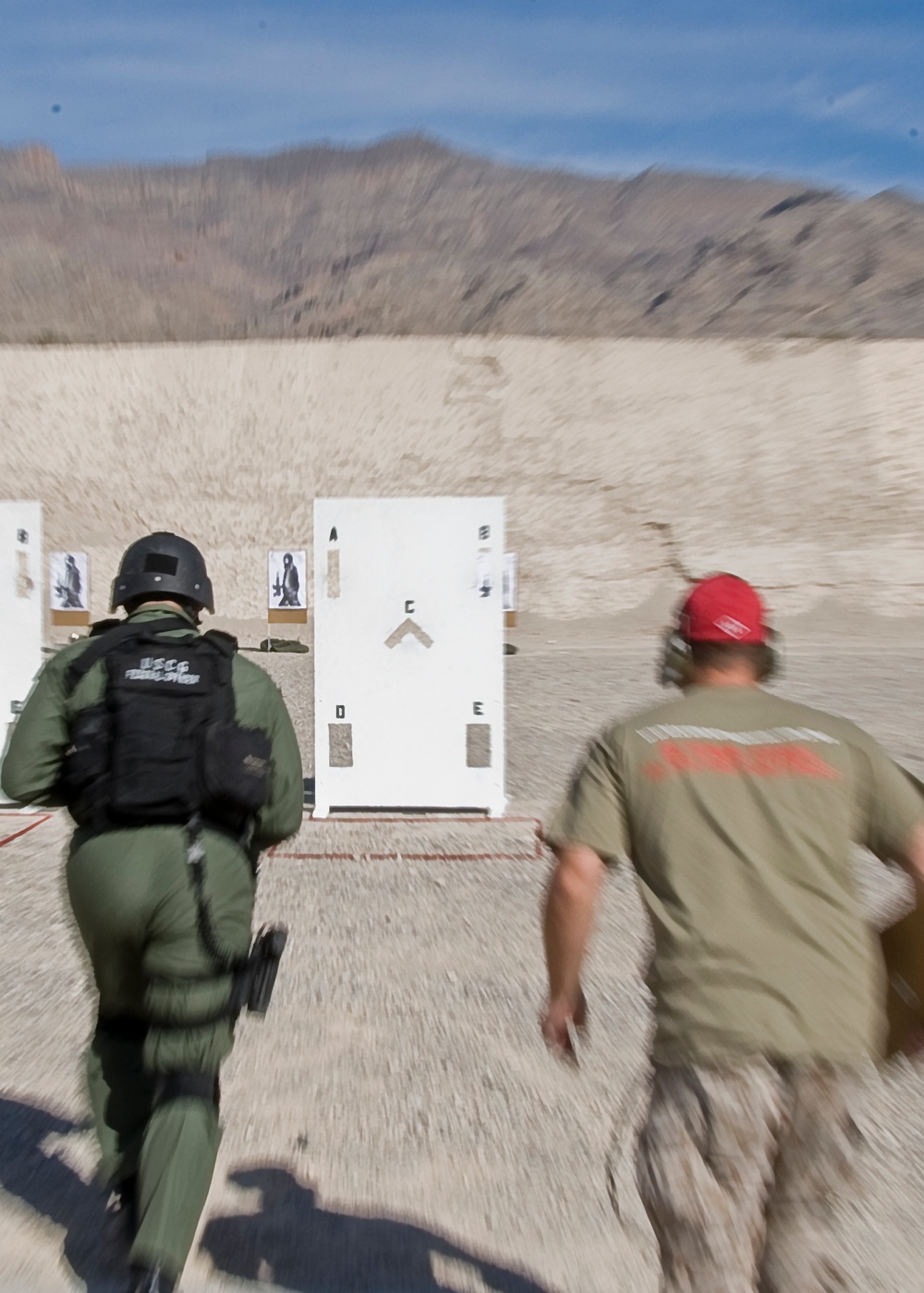 Two individuals are running toward a shooting range training structure in an outdoor desert environment. The person on the left is wearing tactical gear including a helmet and a vest marked 'USCG Federal Officer', while the person on the right wears a red cap and olive/tan clothing. The scene depicts an active law enforcement or military firearms training exercise with paper targets and a white door-frame structure visible downrange.