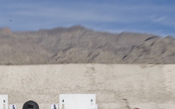 Two individuals are running toward a shooting range training structure in an outdoor desert environment. The person on the left is wearing tactical gear including a helmet and a vest marked 'USCG Federal Officer', while the person on the right wears a red cap and olive/tan clothing. The scene depicts an active law enforcement or military firearms training exercise with paper targets and a white door-frame structure visible downrange.